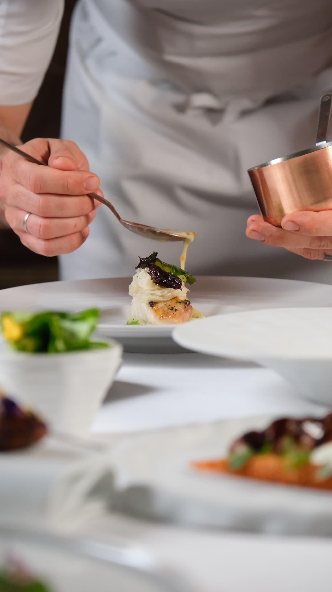 A spoon finishing a fish course with sauce, chef's hands over a white plate