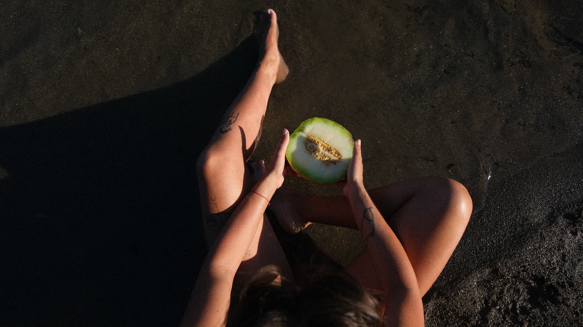 Sunlit legs and hands holding half a melon on black sand