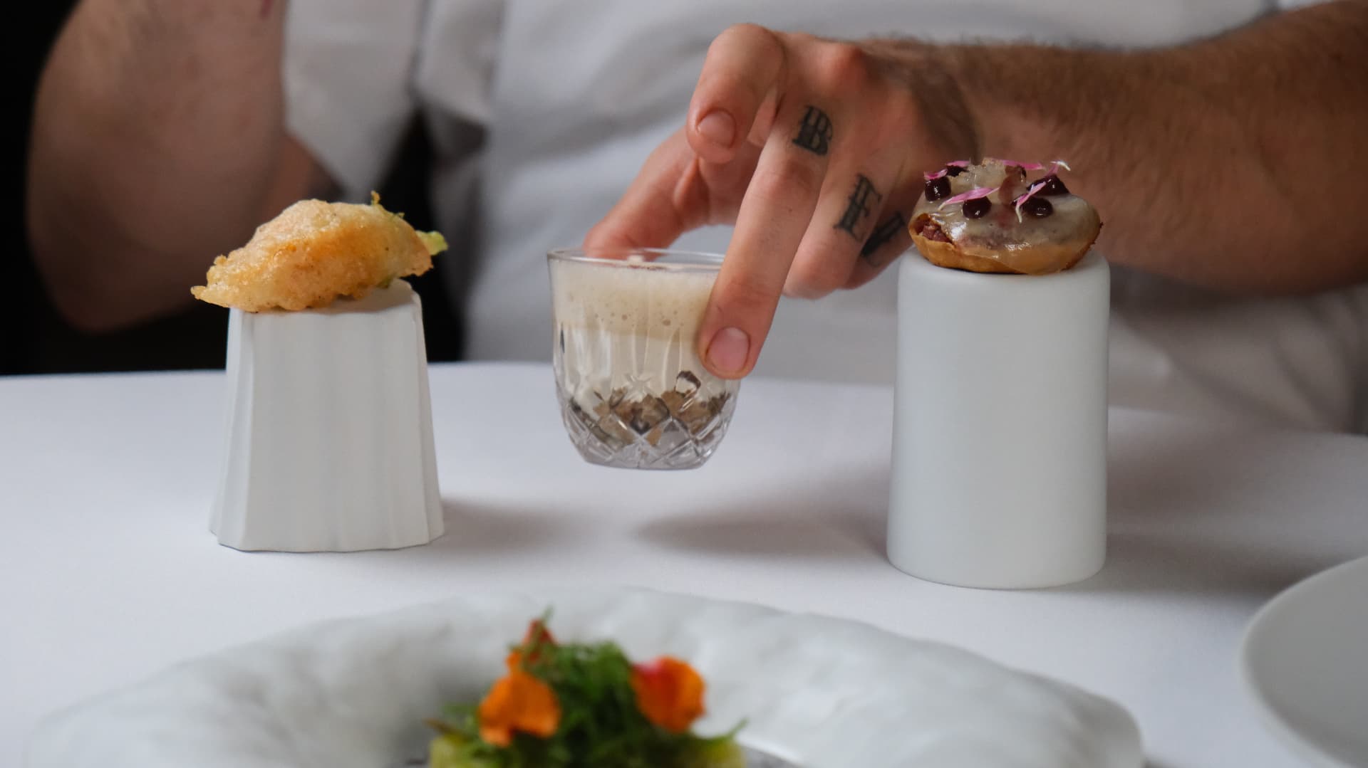 Three canapés on white pedestals, a tattooed hand lifting a cut-glass shot