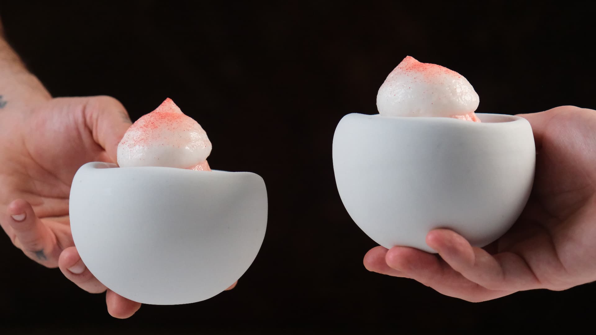 Two hands holding white ceramic bowls with dusted quenelles against black
