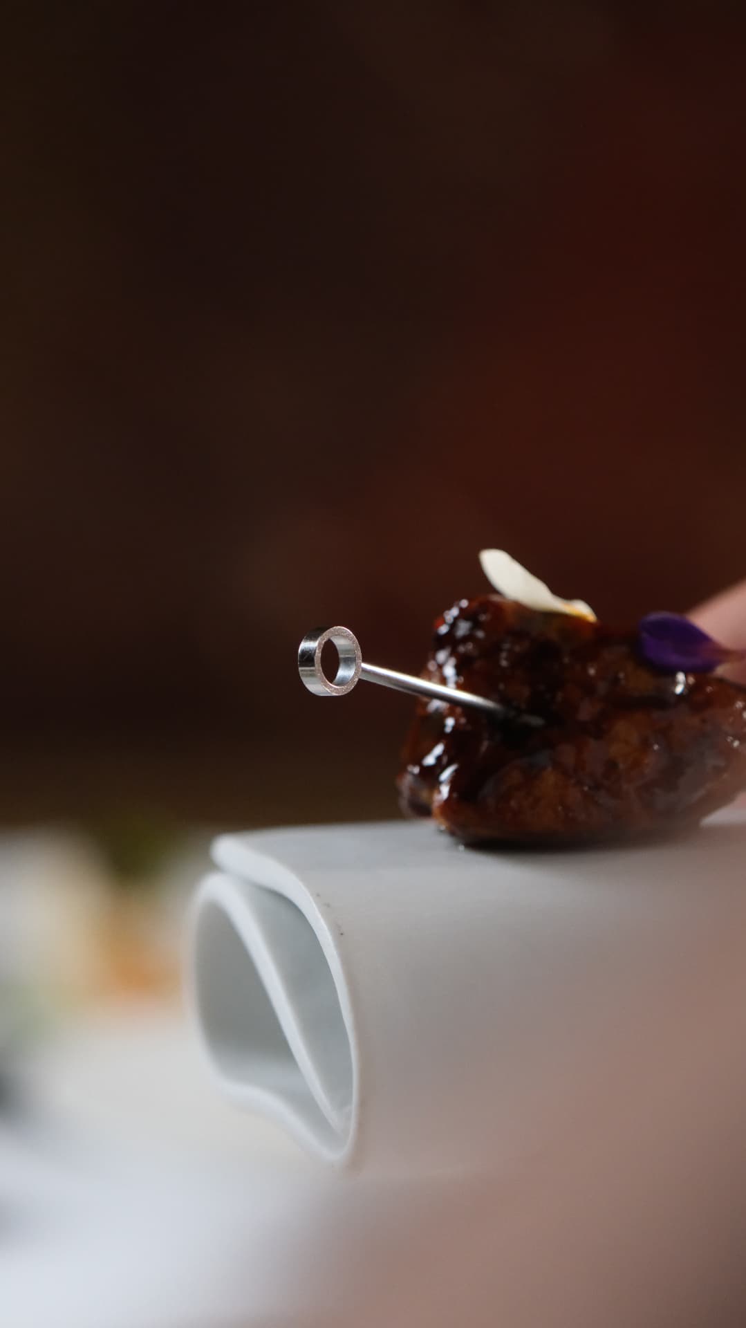 A glazed bite on a steel skewer above a white ceramic form, dark background