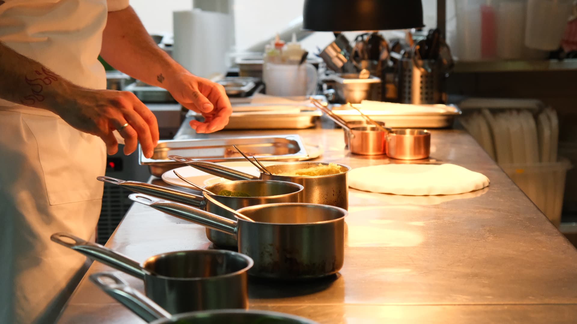 Chef's hands over copper and steel pans lined along a warm-lit kitchen pass
