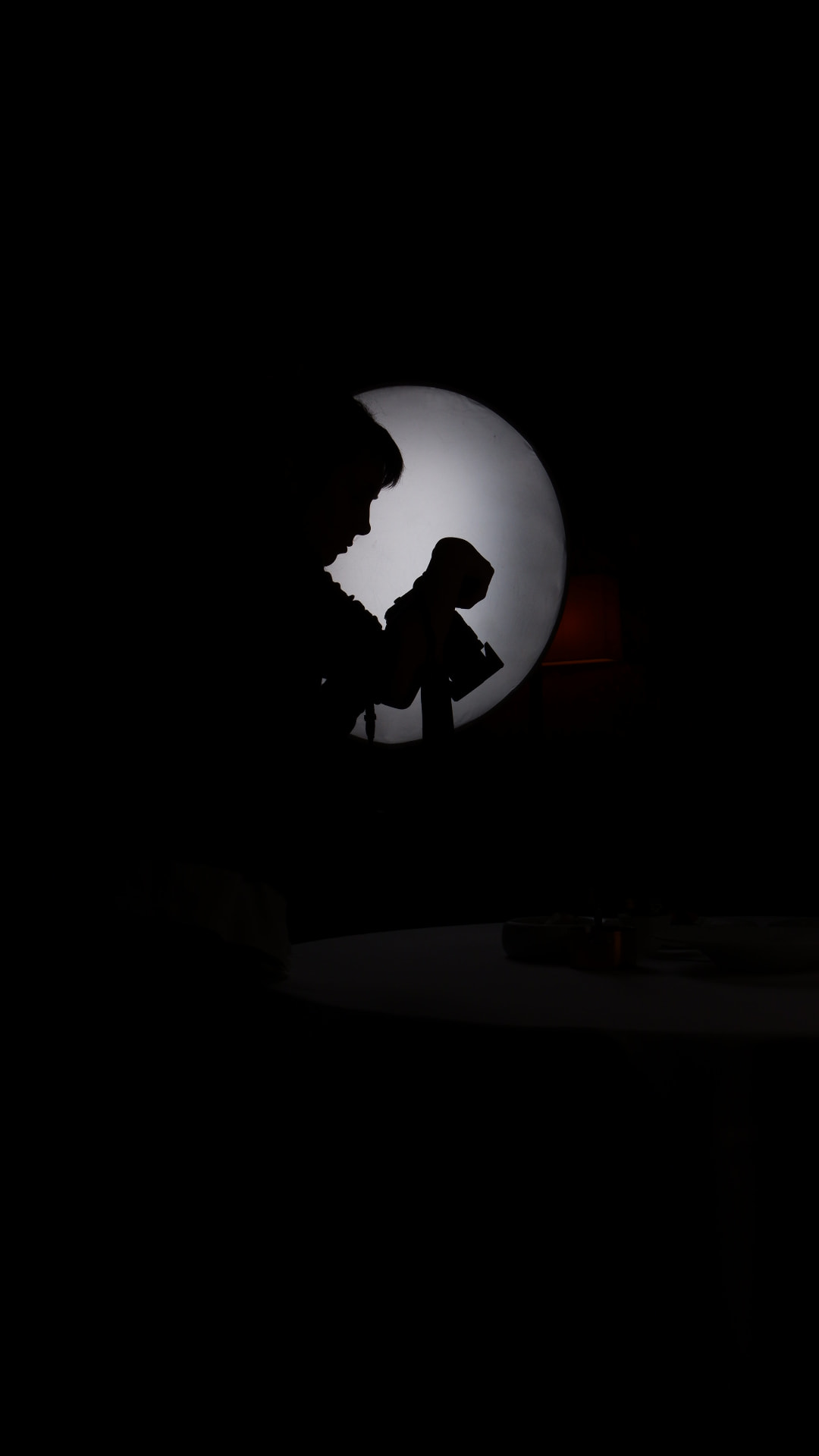 A silhouette against a single round lamp in a nearly dark dining room